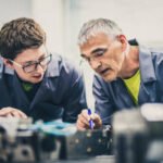 Senior engineer mentoring his student by explaining the functioning of a machine and pointing to its parts.